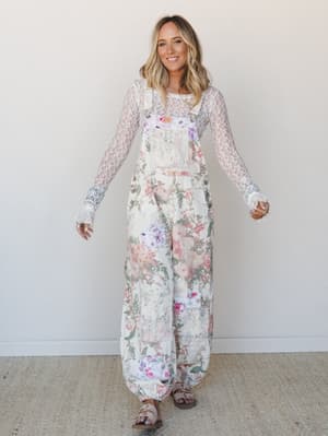 A wide-leg floral overall jumpsuit in cream with soft watercolor blooms is layered over a sheer lace long-sleeve top — the kind of outfit that feels like it was pulled together effortlessly but definitely wasn't.