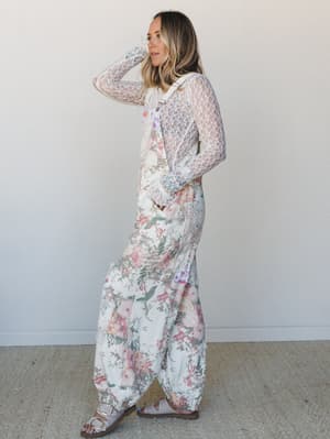 A full-length floral wide-leg jumpsuit layered over a sheer lace long-sleeve top — the kind of outfit that looks effortless but was absolutely thought about, styled with simple sandals against a clean studio wall.