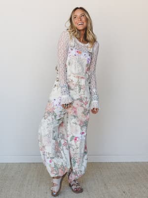 A wide-leg floral overall jumpsuit layered over a sheer lace long-sleeve top, styled with strappy sandals — the whole look has that effortless, garden-party-meets-weekend-wanderer energy.