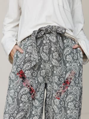A close-up mid-shot of paisley-printed wide-leg pants in washed charcoal and cream, styled with a relaxed white long-sleeve top — the contrast-fabric patch pockets in floral red and blush are the kind of detail you notice on second look.