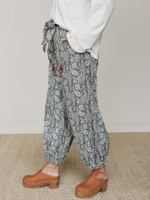 A side-profile shot of relaxed paisley harem pants in charcoal and cream, styled casually with a white pullover, a floral scarf tied at the waist, and warm brown wood-studded clogs — the kind of outfit that feels intentional without trying too hard.