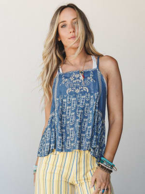 A flowy indigo printed cami top with floral embroidery at the chest and a tiered ruffle hem, styled with yellow stripe pants and layered boho jewelry for a relaxed, sun-soaked summer feel.