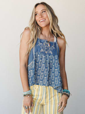 A sun-kissed model smiles off-camera wearing a flowy indigo block-print tank with chest embroidery paired with yellow striped pants and stacked turquoise bracelets — the kind of outfit that feels like a really good summer day.