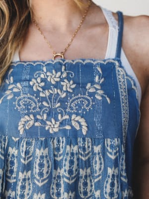 A close-up of an indigo blue top with intricate cream floral embroidery on the bodice and a printed paisley skirt panel below, layered over a white lace-strap top and finished with a delicate gold chain necklace.