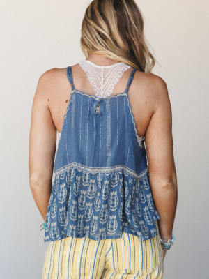 A back-facing shot showing a flowy indigo cami top with a block-print floral hem ruffle, lace racerback inset, and vertical embroidered stripe detail, paired with yellow striped pants — the kind of outfit that feels like a farmers market on a good-we