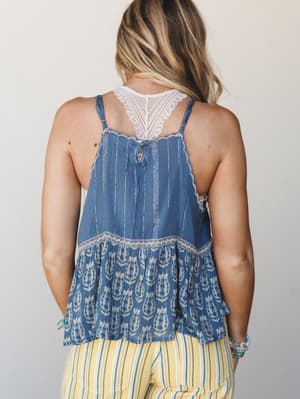 A back-facing shot showing a flowy indigo cami top with a block-print floral hem ruffle, lace racerback inset, and vertical embroidered stripe detail, paired with yellow striped pants — the kind of outfit that feels like a farmers market on a good-we