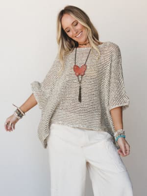 An open-knit poncho in warm natural tones worn over cream wide-leg pants, styled with layered bracelets and a statement pendant necklace — easy, expressive, and the kind of outfit that looks like it came together effortlessly.