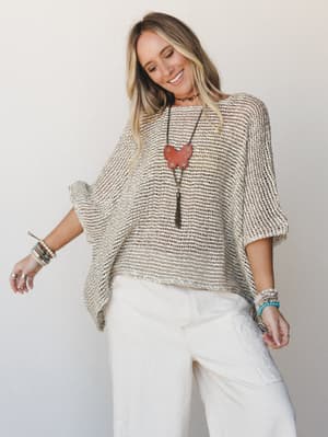 An open-knit poncho in warm natural tones worn over cream wide-leg pants, styled with layered bracelets and a statement pendant necklace — easy, expressive, and the kind of outfit that looks like it came together effortlessly.