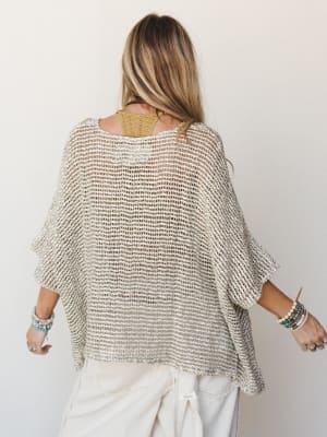 Shot from behind, a model wears an oversized open-knit poncho in natural cream and tan over a golden lace bralette and white wide-leg pants, her wrists stacked with beaded and turquoise bracelets — effortlessly relaxed and very much herself.