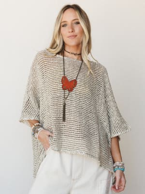 An oversized open-knit poncho in natural cream and tan tones styled over white wide-leg pants, layered with a statement butterfly necklace and stacked bracelets — effortlessly relaxed with a collected, boho edge.
