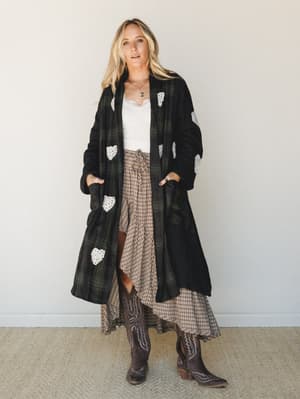 A long plaid duster jacket with whimsical crochet heart appliqués is layered over a lace-trim cami and flowing plaid midi skirt, grounded by dark cowboy boots — the kind of outfit that looks like it came together effortlessly but definitely didn't.