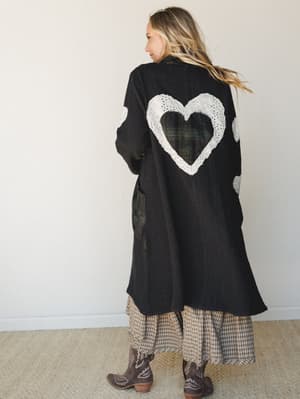 A model faces away showing the dramatic back of a long black textured jacket featuring cut-out heart appliqués trimmed in white eyelet lace, layered over a tan plaid maxi skirt and paired with dark brown cowboy boots — equal parts artsy and lived-in.
