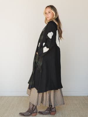A long black knit duster jacket with eyelet lace heart appliqués steals the show here — layered over a tan plaid midi skirt and dark green plaid panel, then grounded with worn-in western ankle boots for a look that feels both intentional and effortle