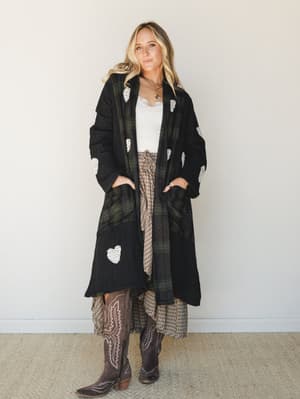 A long black plaid duster jacket with crochet heart appliqués is layered over a lace-trim cami and a flowy plaid midi skirt, finished with tall brown cowboy boots — the kind of outfit that feels collected without trying.