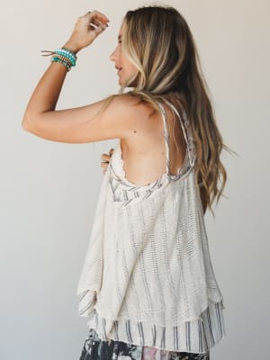 Shot from behind, a model shows off a flowy open-back knit tank in natural cream layered over a striped camisole, styled with a floral skirt and stacked turquoise bracelets — easy, sun-warmed, and effortlessly put-together.