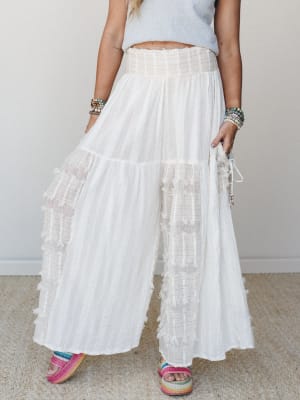 Flowy white wide-leg pants with tiered smocked waist and fringe patchwork detailing, styled with a cropped grey top, stacked bead bracelets, and colorful woven platform sandals — effortless and a little dreamy.