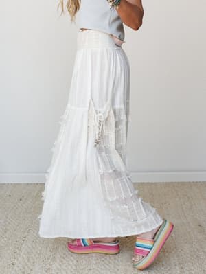 A flowy white wide-leg pant with tiered lace and fringe detailing is styled with a cropped grey tee and rainbow platform espadrilles — effortlessly boho with just enough whimsy.