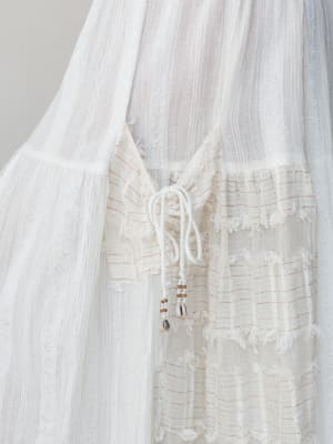 A close-up detail shot of a white wide-leg pant in a sheer gauze fabric, highlighting a drawstring tie closure with cowrie shell and seed bead embellishments against mixed texture panels with subtle red stitch striping.