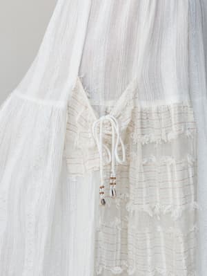 A close-up detail shot of a white wide-leg pant in a sheer gauze fabric, highlighting a drawstring tie closure with cowrie shell and seed bead embellishments against mixed texture panels with subtle red stitch striping.