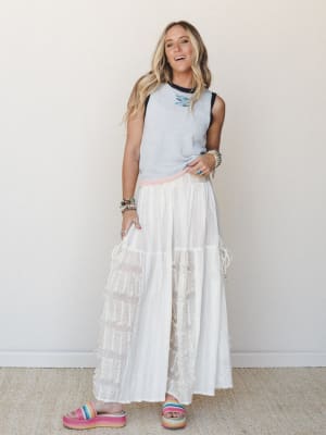 A full-body model shot of a breezy white wide-leg pant with fringe trim tiers, styled with a sleeveless gray sweater vest and rainbow platform sandals against a clean neutral wall — easy, free-spirited, and genuinely fun.