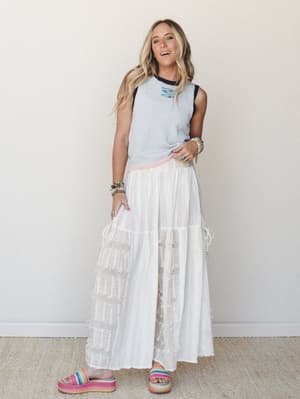 A full-body model shot of a breezy white wide-leg pant with fringe trim tiers, styled with a sleeveless gray sweater vest and rainbow platform sandals against a clean neutral wall — easy, free-spirited, and genuinely fun.