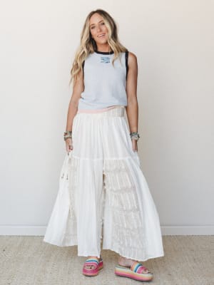 A breezy full-body shot of a model in flowing white wide-leg pants with fringe texture details, paired with a light blue contrast-trim muscle tank and rainbow platform sandals — casual and free-spirited with a summer-boho feel.