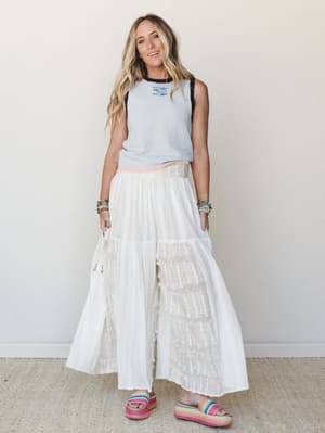 A breezy full-body shot of a model in flowing white wide-leg pants with fringe texture details, paired with a light blue contrast-trim muscle tank and rainbow platform sandals — casual and free-spirited with a summer-boho feel.