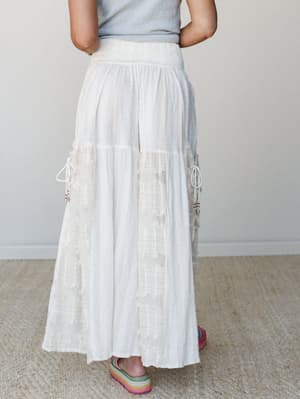 A flowy white wide-leg pant with tiered paneling, fringe trim, and cord tie details styled with a grey top and rainbow platform sandals against a soft neutral studio backdrop — effortlessly boho with a festival-ready ease.