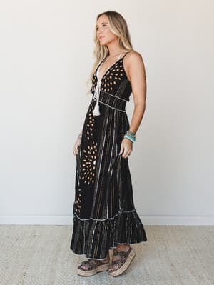 A black maxi dress with an abstract mixed print — oval motifs and vertical stripe details — styled with stacked turquoise bracelets and platform espadrilles for a free-spirited, warm-weather feel.