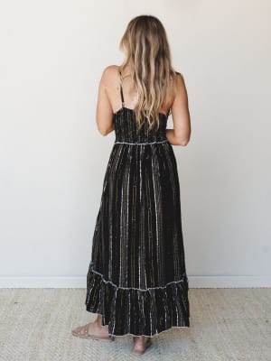 A back-facing shot of a smocked maxi dress in black with metallic stripe detailing — the kind of dress that works just as hard at a summer wedding as it does at a rooftop dinner.