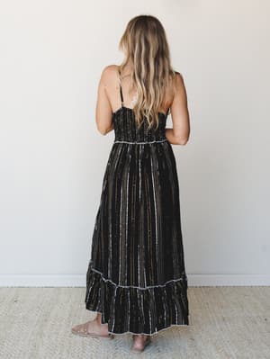 A back-facing shot of a smocked maxi dress in black with metallic stripe detailing — the kind of dress that works just as hard at a summer wedding as it does at a rooftop dinner.