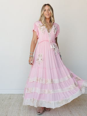 A breezy pink maxi dress with floral appliqué and tiered lace-trimmed skirt twirls mid-movement, styled with layered jewelry and sandals against a soft studio wall — the kind of dress that makes you feel like you're already somewhere warm.
