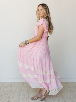 A pink maxi dress with flutter sleeves and lace-trimmed tiered skirt catches mid-twirl, styled with turquoise stacked bracelets and pearl-detail sandals — soft, romantic, and ready for something worth remembering.