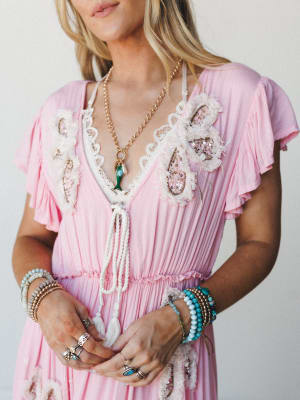 A soft pink maxi dress with lace and floral appliqué neckline detail, styled with layered beaded bracelets, stacked rings, and a gold charm necklace — the kind of outfit that feels equal parts romantic and collected.