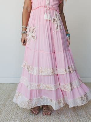 A dreamy pink maxi dress with tiered ruffles, floral appliqués, and embroidered lace trim bands styled with layered beaded bracelets and flat sandals — the kind of dress that makes you want somewhere worthy to wear it.