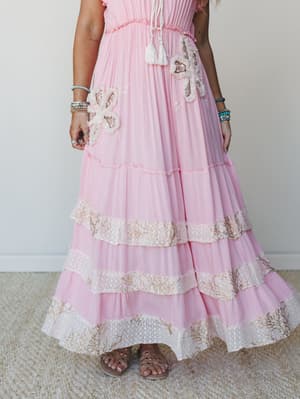 A dreamy pink maxi dress with tiered ruffles, floral appliqués, and embroidered lace trim bands styled with layered beaded bracelets and flat sandals — the kind of dress that makes you want somewhere worthy to wear it.