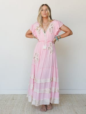 A breezy pink maxi dress with floral appliqué detailing and tassel tie neckline, styled with layered boho jewelry — the kind of dress that makes you want to book a trip somewhere warm.