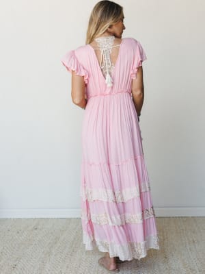 A breezy pink maxi dress shot from behind, showing off its open lace back with tassel tie, flutter sleeves, and tiered lace-trimmed skirt — the kind of dress that makes you want to spin around at a garden party.