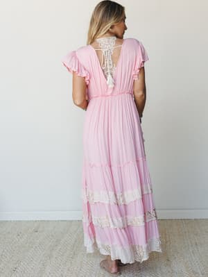 A breezy pink maxi dress shot from behind, showing off its open lace back with tassel tie, flutter sleeves, and tiered lace-trimmed skirt — the kind of dress that makes you want to spin around at a garden party.