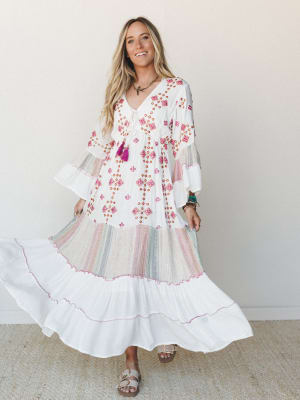 A flowing ivory maxi dress with colorful embroidery and tiered ruffles twirls in motion on a smiling model — the kind of dress that makes you want to spin in a field or wander a farmers market with coffee in hand.