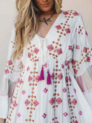 A close-up of an ivory maxi dress covered in geometric embroidery and mirror embellishments in magenta and rust, with bell sleeves, a lace-up neckline, and magenta tassel ties — the kind of dress that makes you feel like you packed perfectly for a tr