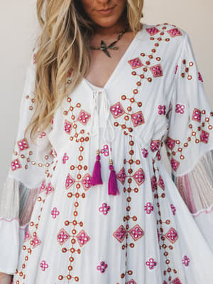 A close-up of an ivory maxi dress covered in geometric embroidery and mirror embellishments in magenta and rust, with bell sleeves, a lace-up neckline, and magenta tassel ties — the kind of dress that makes you feel like you packed perfectly for a tr