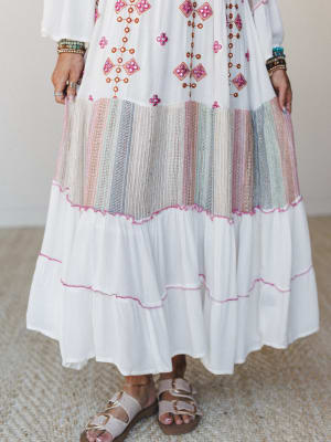 A close-up of a flowing white maxi dress with colorful geometric embroidery and mirror embellishments on the bodice, a multicolor woven stripe inset tier, and soft pink stitch trim along the tiered hem — styled with stacked bracelets and buckle sanda