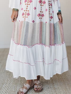 A close-up of a flowing white maxi dress with colorful geometric embroidery and mirror embellishments on the bodice, a multicolor woven stripe inset tier, and soft pink stitch trim along the tiered hem — styled with stacked bracelets and buckle sanda