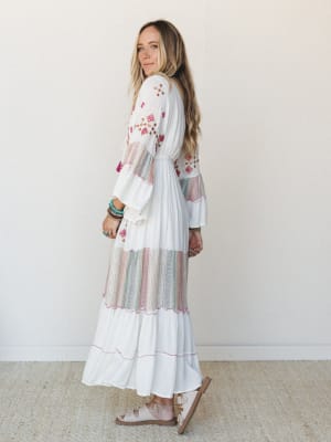 A white embroidered maxi dress with pops of pink and multicolor detail, styled with stacked turquoise bracelets and flat sandals — the kind of dress that looks equally at home at a beach wedding or a Saturday farmers market.