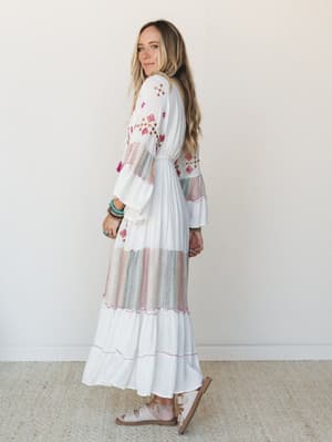 A white embroidered maxi dress with pops of pink and multicolor detail, styled with stacked turquoise bracelets and flat sandals — the kind of dress that looks equally at home at a beach wedding or a Saturday farmers market.