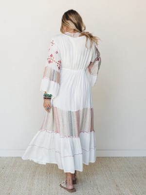 A white embroidered maxi dress shot from behind, with tiered skirt panels, floral embroidery on the sleeves, and soft mixed-stripe sheer insets — styled with turquoise stacked bracelets and tan sandals for an easy, expressive boho feel.