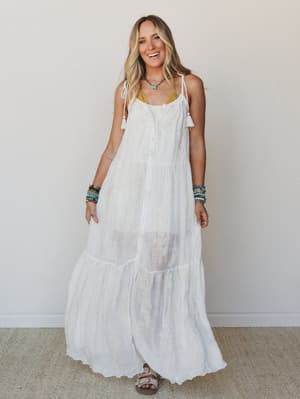 A white tiered maxi dress with delicate lace trim and tassel-tie straps, styled with stacked beaded bracelets and a turquoise pendant necklace — the kind of dress that looks equally at home on a beach boardwalk or a backyard wedding.