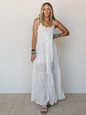 A white embroidered maxi dress with a breezy, tiered silhouette styled with stacked beaded bracelets and a turquoise necklace — the kind of outfit that looks equally at home on a beach boardwalk or at a backyard wedding.