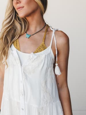 A close-up lifestyle shot of a white embroidered maxi dress layered over a mustard lace bralette, styled with a turquoise butterfly pendant necklace — the kind of effortless boho layering that looks intentional without trying too hard.